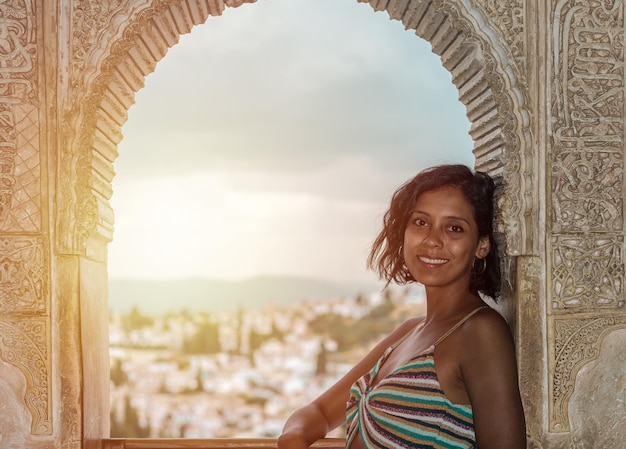 Hermosa mujer viajera posando en una ventana decorada con elementos geométricos del arte islámico a través de la cual se puede ver un pequeño pueblo de casas blancas