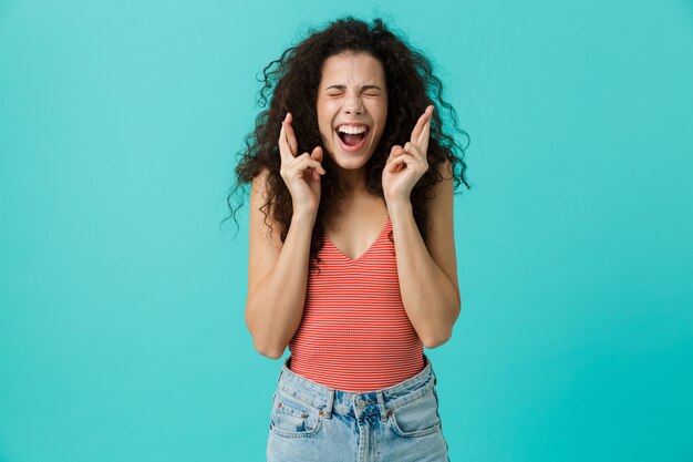 Foto hermosa mujer vistiendo ropa casual gritando y manteniendo los dedos cruzados, de pie aislado sobre la pared azul