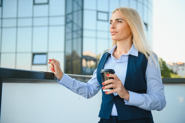 Hermosa mujer yendo a trabajar con café caminando cerca del edificio de oficinas Retrato de mujer de negocios exitosa sosteniendo una taza de bebida caliente