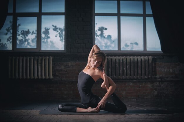 Hermosa mujer de yogini en forma deportiva practica yoga asana Eka Pada Rajakapotasana en el pasillo oscuro