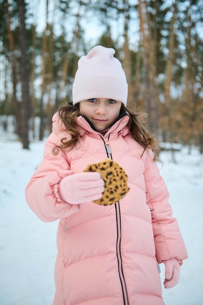 Hermosa niña de 4 años, adorable niña europea con ropa de invierno cálida rosa pastel sosteniendo unas deliciosas galletas comiendo al aire libre en una naturaleza cubierta de nieve Disfruta de unas maravillosas vacaciones de invierno