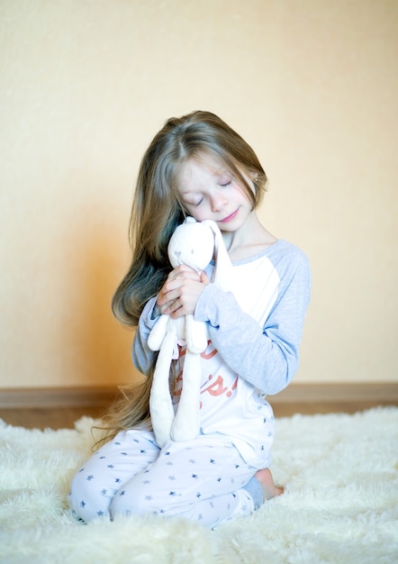 Hermosa niña de 8 años con largo cabello rubio y ojos azules se sienta en pijama sobre una alfombra mullida en la habitación y abraza suavemente a un conejito de juguete