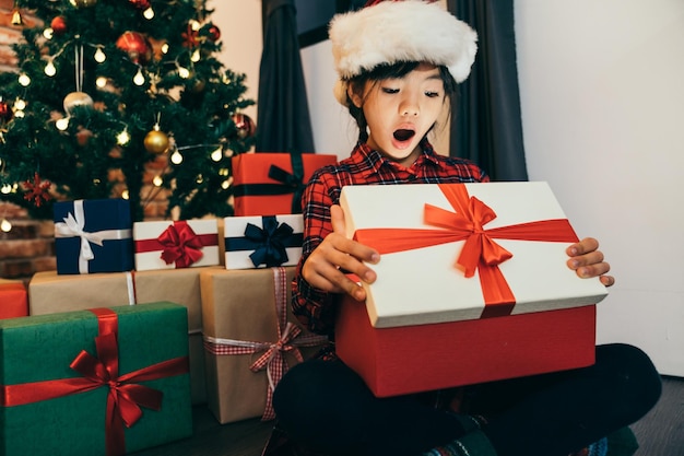 hermosa niña abre su regalo de navidad en el día del boxeo. un montón de regalos de Navidad para ella en segundo plano. árbol decorado con bolas de colores en el salón.