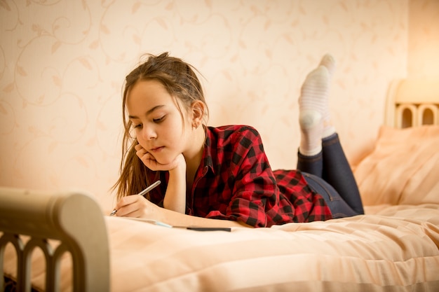 Hermosa niña adolescente sonriente acostado en la cama y escribiendo en el diario