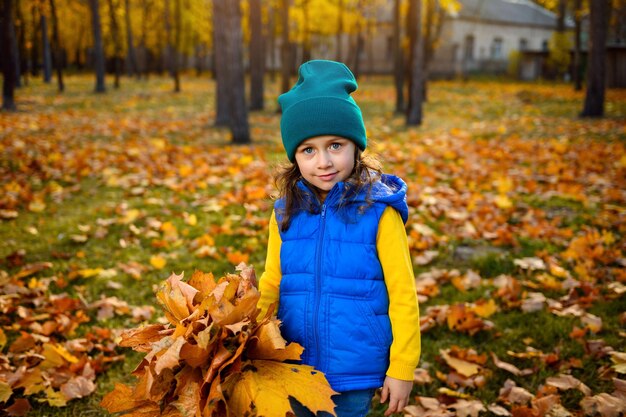 Hermosa niña adorable niña en ropa de abrigo de colores brillantes con ramo recogido de hojas de otoño de pie contra el fondo del parque dorado de otoño al atardecer con rayos de sol cayendo