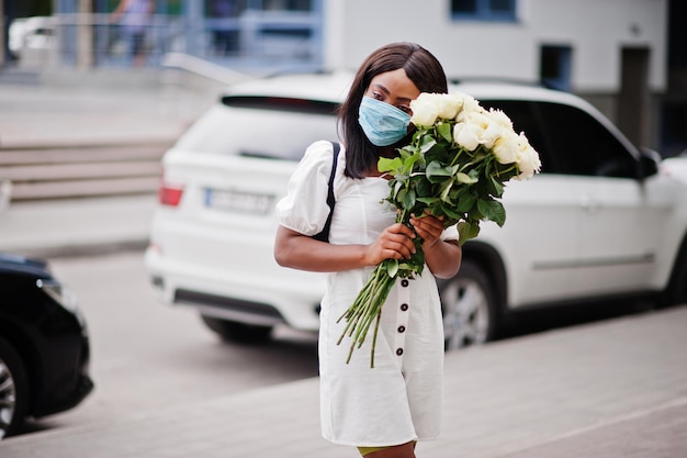 Hermosa niña afroamericana con ramo de flores de rosas blancas usa mascarilla durante la cuarentena del coronavirus.