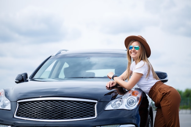 Una hermosa niña de apariencia europea con gafas y un sombrero marrón se encuentra cerca de un automóvil negro. Sesión de fotos cerca del coche.