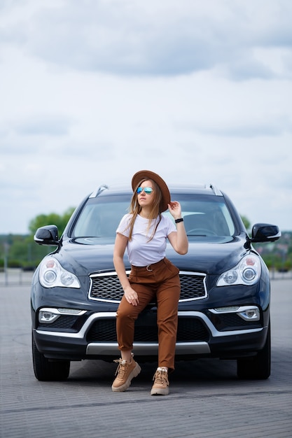 Una hermosa niña de apariencia europea con gafas y un sombrero marrón está parada cerca de un automóvil negro. Foto cerca del auto.