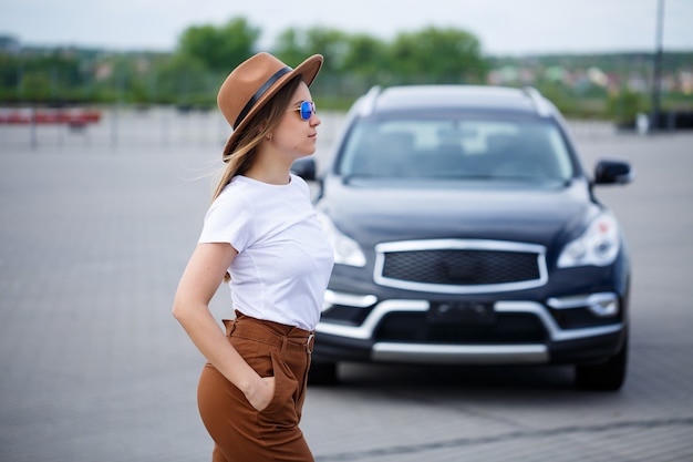 Una hermosa niña de apariencia europea con gafas y un sombrero marrón está parada cerca de un automóvil negro.