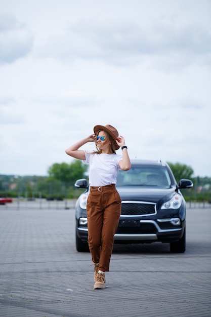 Una hermosa niña de apariencia europea con gafas y un sombrero marrón está parada cerca de un automóvil negro.