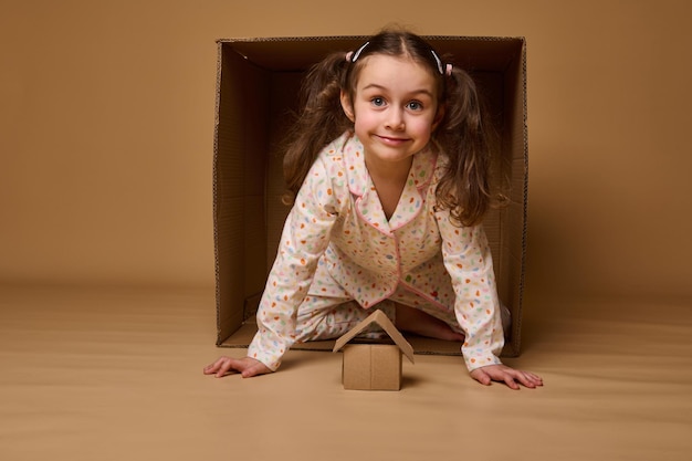 Hermosa niña asomándose desde detrás de una caja de cartón mirando a la cámara Caja de cartón artesanal sobre un fondo beige con espacio de copia para anuncios Seguro de vivienda y concepto de bienes raíces