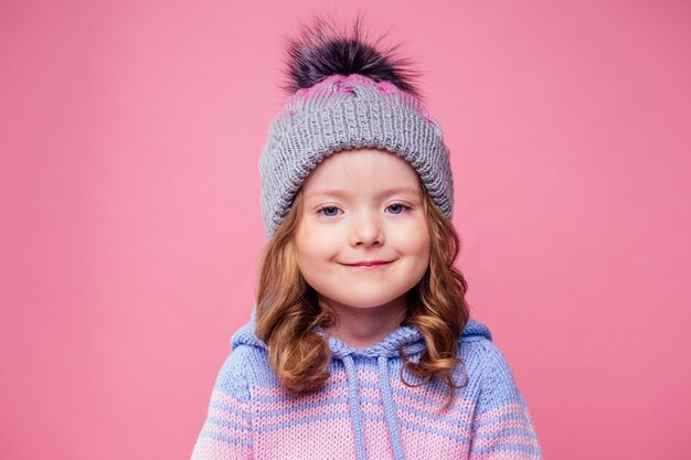 Hermosa niña de blone en el gorro de invierno y bufanda sobre fondo rosa en el estudio. Mirada de niño de moda con estilo de otoño.