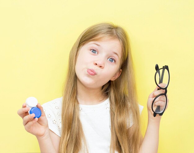 Una hermosa niña de cabello largo sostiene un recipiente con lentes y gafas en sus manos. El concepto de problemas de visión en los niños. Fondo amarillo.