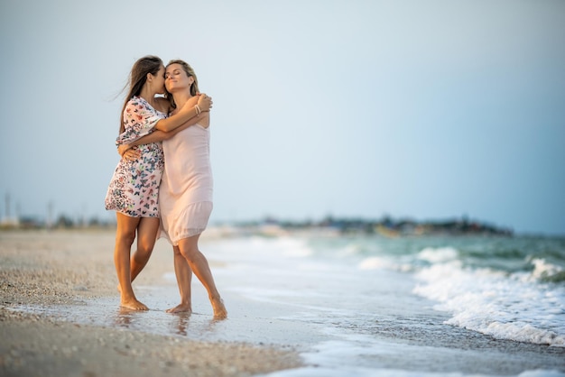 Una hermosa niña de cabello oscuro bronceado con un vestido floral brillante abraza a su madre feliz, cariñosa y de cabello rubio con un delicado vestido rosa mientras camina por la playa de arena cerca del mar con olas espumosas