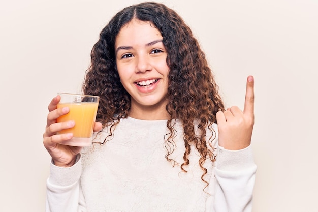 Hermosa niña con cabello rizado bebiendo un vaso de jugo de naranja sonriendo feliz señalando con la mano y el dedo al costado