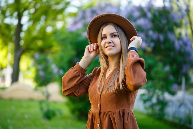 Una hermosa niña de cabello rubio sostiene un sombrero en sus manos y se encuentra cerca de un arbusto de lilas. Mujer joven en el jardín con árboles en flor