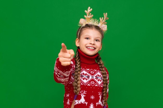 Una hermosa niña con un cálido suéter festivo estira los brazos hacia adelante y sonríe. El concepto de Navidad es una niña con cuernos de Navidad.