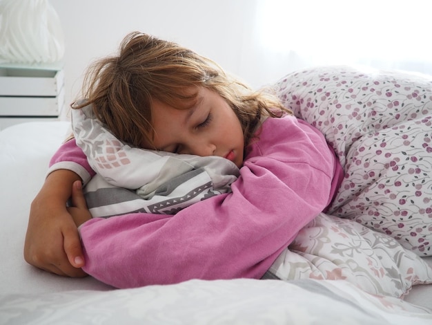 Una hermosa niña caucásica de 8 años con cabello rubio vestido con pijama rosa duerme en una cama con una manta mullida abrazando una almohada La luz suave del sol de la mañana se filtra por la ventana
