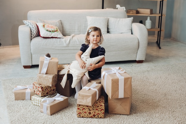 Una hermosa niña caucásica con cabello ondulado rubio corto con un lindo vestido negro se sienta junto a los regalos de Navidad en una gran sala de luz de la casa