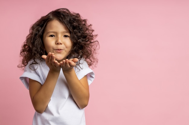 Hermosa niña caucásica con peinado rizado en camiseta blanca, soplando beso de aire sobre las palmas, expresando amor, aislado