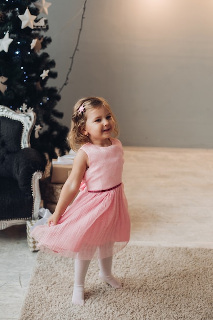 Una hermosa niña caucásica con el pelo corto y ondulado con un lindo vestido rosa se regocija cerca del árbol de Navidad en una gran sala de luz de la casa