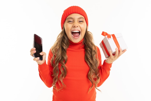 Hermosa niña caucásica sostiene una caja blanca con regalo en una mano y un teléfono inteligente en la otra mano, sonríe y elige entre ella aislado en blanco