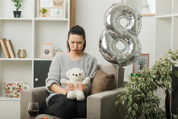 Foto hermosa niña con el ceño fruncido en el día de la mujer feliz sosteniendo un oso de peluche sentado en un sillón en la sala de estar