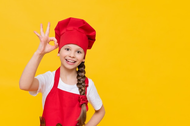 Una hermosa niña cocinera en un traje para la cocina muestra una señal de mano de que todo está bien Una niña chef en un fondo aislado el concepto de cómo los niños cocinan comida en casa