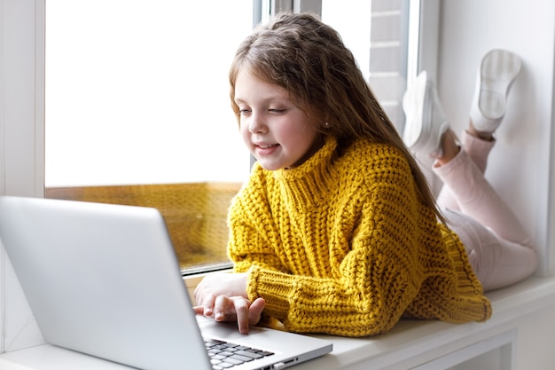 Una hermosa niña con una computadora portátil en casa en la ventana mira la pantalla y sonríe