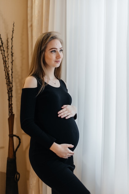 Una hermosa niña embarazada está de pie en una habitación cerca de la ventana.