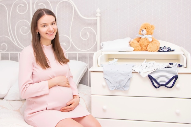 Una hermosa niña embarazada está sentada en la cama y sus manos están sobre su estómago. Sobre la mesa para cambiar pañales hay cosas para el recién nacido.