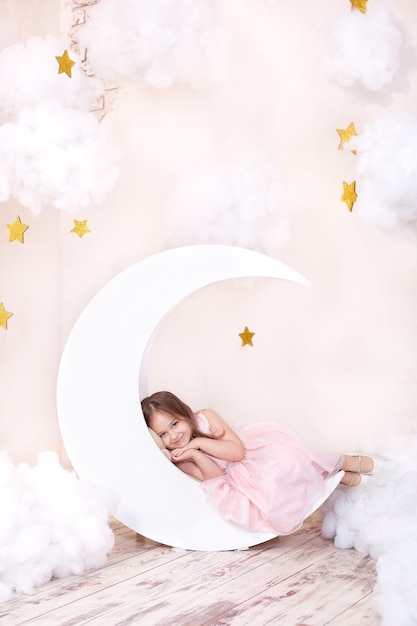 Hermosa niña se encuentra en estudio con decoración de luna, estrellas y nubes. El niño está soñando. Niña linda se sienta en una luna decorativa con nubes de algodón y estrellas. Concepto de sueño saludable Infancia