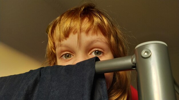 Una hermosa niña se esconde detrás del respaldo de una litera en la que cuelga una tela de terciopelo azul oscuro. El niño tiene ojos grises y cabello rubio. La mitad superior de la cara es visible Esconder y buscar