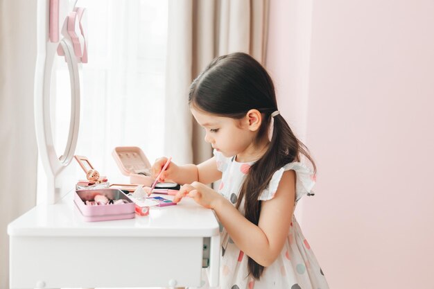 Una hermosa niña en el espejo acicalándose una niña está sentada en una mesa para niños y está usando cosméticos para niños