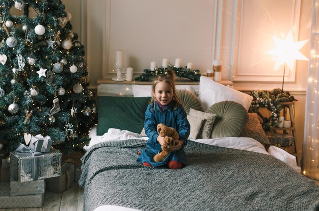 Una hermosa niña está sentada en la cama abrazando un peluche cerca de un árbol de Navidad decorado