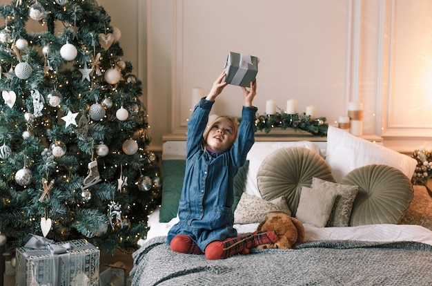 Una hermosa niña está sentada en la cama junto al árbol de Navidad sosteniendo un regalo en lo alto