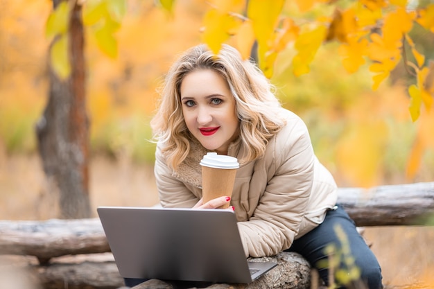 Una hermosa niña está sentada frente a una computadora portátil en un parque de otoño con café en la mano