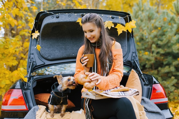 Una hermosa niña está sentada en el maletero de un auto negro con su perro sonriendo bebiendo té en el almuerzo admirando la naturaleza del bosque y disfrutando de la libertad