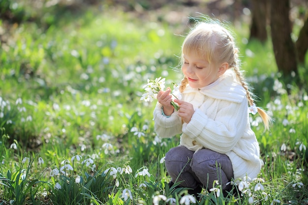 Una hermosa niña está sentada en un prado de flores. Una niña con un suéter de punto blanco está considerando una campanilla de invierno. Tiempo de Pascua. Bosque soleado de primavera