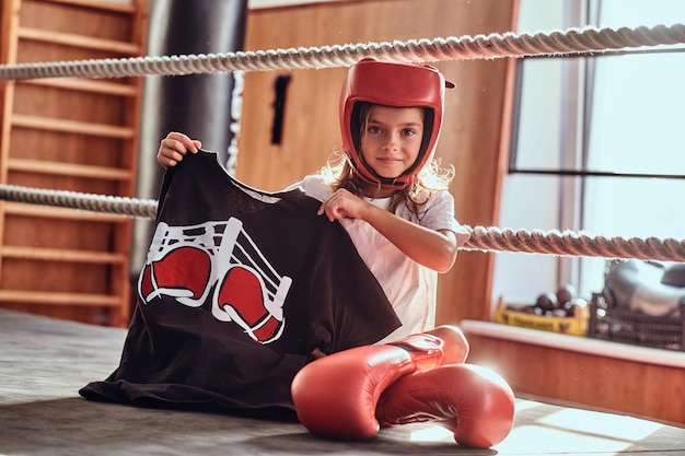 Hermosa niña está sentada en el ring de boxeo con uniforme de boxeador, guantes y casco.