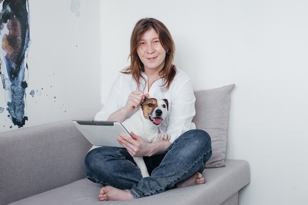 La hermosa niña está sentada en el sofá de su casa con una tableta en sus manos, junto a un perro Jack Russell