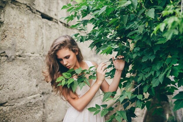 Hermosa niña feliz con cabello rizado natural en vestido blanco cerca de hojas de árbol verde