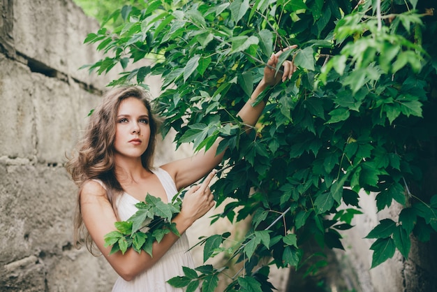 Hermosa niña feliz con cabello rizado natural en vestido blanco cerca de hojas de árbol verde