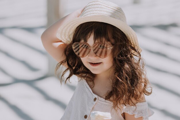 Hermosa niña con gafas de sol vestido blanco y sombrero comiendo helado en la playa en verano