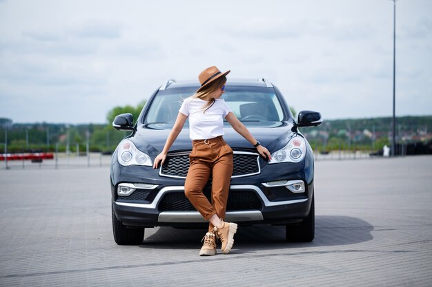 Una hermosa niña con gafas y un sombrero marrón está parada cerca de un auto negro. Mujer joven con coche en el estacionamiento