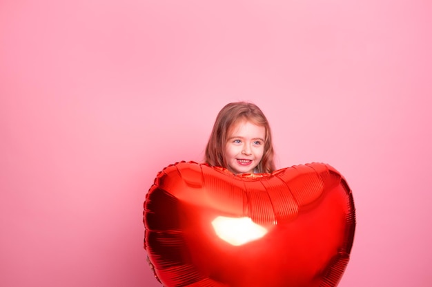 Hermosa niña con globo de corazón rojo sobre fondo rosa celebrando el día de san valentín