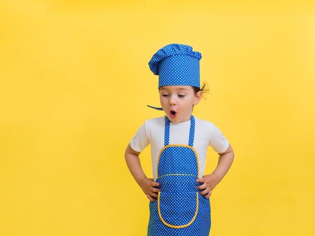 Hermosa niña con gorro de cocinero y delantal aislado en el espacio amarillo. La niña mira hacia abajo y se sorprende