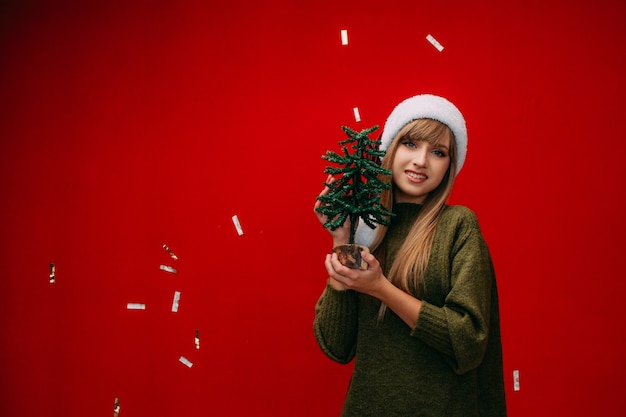 Una hermosa niña con un gorro de Papá Noel tiene un pequeño árbol de Navidad en sus manos sobre un fondo rojo.