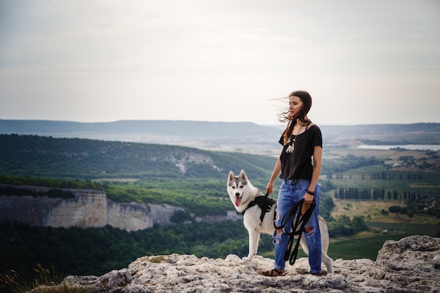 Hermosa niña juega con un perro, husky gris y blanco, en las montañas al atardecer. Niña india y su lobo
