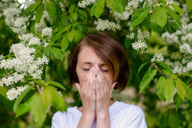 Una hermosa niña se para junto a la cereza de pájaro floreciente y estornuda. Ella sufre de alergia a la floración en primavera.